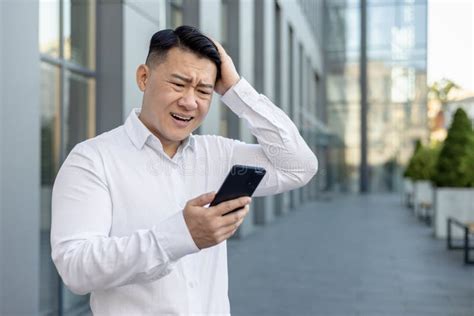 Upset Young Asian Man Standing Near an Office Building, Looking Frustrated at the Phone Screen ...